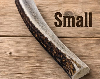 May include: A small, natural antler chew for dogs, with the word "Small" in brown text. The antler has a light gray and brown color, with a textured surface. The background is a wooden surface.