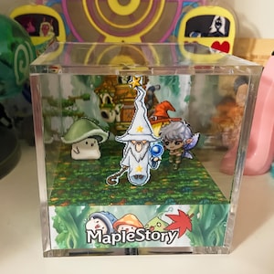 Puede incluir: Una caja de presentación de cubo de acrílico transparente con un diorama en el interior que representa personajes del videojuego MapleStory. El diorama incluye un mago, un hongo y un personaje pequeño con un bastón. La base del diorama es un paisaje verde y marrón con las palabras "MapleStory" impresas en él.