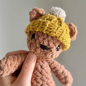 Puede incluir: Un oso de ganchillo hecho a mano en tonos marrones, con un sombrero amarillo con un pompón blanco. El oso tiene ojos negros y una boca negra cosida. El peluche se sostiene en una mano sobre un fondo neutro.