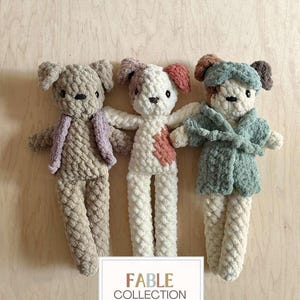 May include: Three handmade crochet dog dolls in neutral colors. One doll wears a lavender vest, another has a pink heart, and the third wears a teal robe. The dolls have floppy ears and are standing in front of a wooden background. The text "FABLE COLLECTION" is at the bottom.