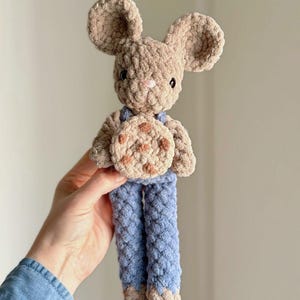 Könnte beinhalten: Eine handgefertigte Häkelmaus-Puppe mit großen Ohren, die einen Keks hält. Die Maus ist hellbraun mit blauen Latzhosen und einem hellbraunen Keks. Die Puppe ist weich und kuschelig, perfekt für ein Kinderspielzeug oder die Dekoration des Kinderzimmers.