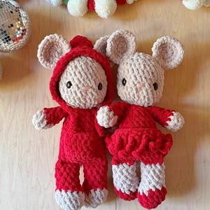 May include: Two handmade crochet plush mice with beige yarn bodies and dark eyes. One mouse wears a red hooded jumpsuit, while the other wears a red dress with a ruffled skirt. Both mice have beige ears and red accents on their feet and hands.