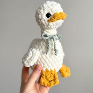 May include: A handmade white crochet duck with a yellow beak and feet. The duck has black eyes and a light blue bow tied around its neck. The plush toy is soft and fluffy, perfect for a child's room or as a decorative item.