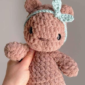 Op de afbeelding: Een gehaakte berenknuffel in oudroze met een zachte, getextureerde garen afwerking. De beer heeft twee kleine ronde oren, donkere plastic veiligheidsogen en een licht mintgroene gehaakte hoofdband versierd met een klein strikje aan de zijkant.