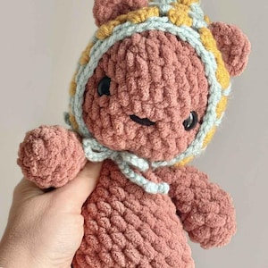 May include: A crocheted plush toy in a dusty rose color, featuring a rounded head with small ears and black bead eyes. The toy wears a bonnet with a yellow and light blue checkered pattern, tied under the chin with light blue yarn strings.