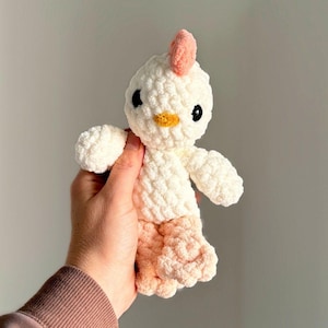 May include: A handmade, plush chicken toy. The body is white with peach-colored feet. It has a peach comb and a yellow beak. The eyes are black. The toy is soft and appears to be crocheted.
