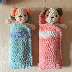 Può includere: Due peluche a forma di cane fatti a mano all'uncinetto, avvolti in coperte accoglienti. Un cane ha il pelo arancione e crema, l'altro marrone e bianco. Le coperte sono azzurre a righe viola e rosa a righe bianche.