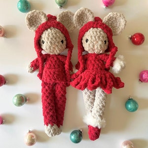 May include: Two crocheted mouse toys wearing red hooded outfits. One mouse is wearing a dress and the other is wearing pants. The toys are surrounded by Christmas ornaments.