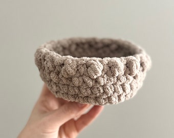May include: A small, round, beige crochet basket with a textured, bobble-like design. The basket is held in a hand, showcasing its soft, plush appearance. This handmade item is ideal for home decor and storage.
