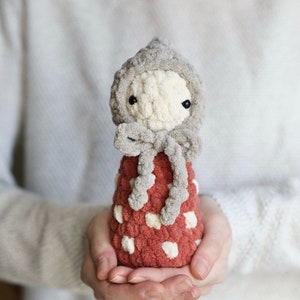 May include: A crocheted doll with a grey hooded hat and a red and white dress. The doll is being held in the hands of a person.
