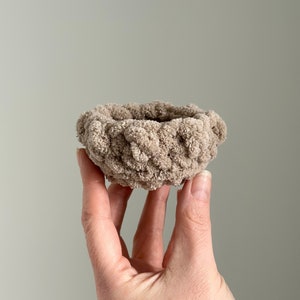 May include: A small, hand-knitted bowl made of beige yarn. The bowl is woven in a basket weave pattern.