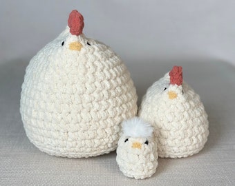 May include: Three crocheted plush chickens in various sizes. The largest chicken is white with a red comb and yellow beak. The medium-sized chicken is white with a red comb and yellow beak. The smallest chicken is white with a white fluffy top and yellow beak.