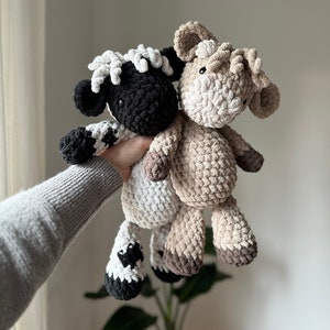 Pode incluir: Dois animais de peluche de crochê, uma ovelha preto e branco e uma vaca marrom, são segurados em uma mão. A ovelha tem uma cara branca e cascos pretos. A vaca tem uma cara marrom e cascos marrons.