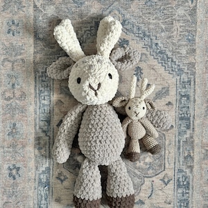 May include: Two crocheted stuffed animals in the shape of goats. The larger goat is grey and white with brown hooves. The smaller goat is beige with brown hooves. The goats are sitting on a patterned rug.
