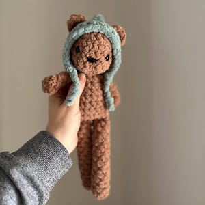 May include: A brown crocheted teddy bear wearing a light blue crocheted hat. The bear has black eyes and a brown nose.