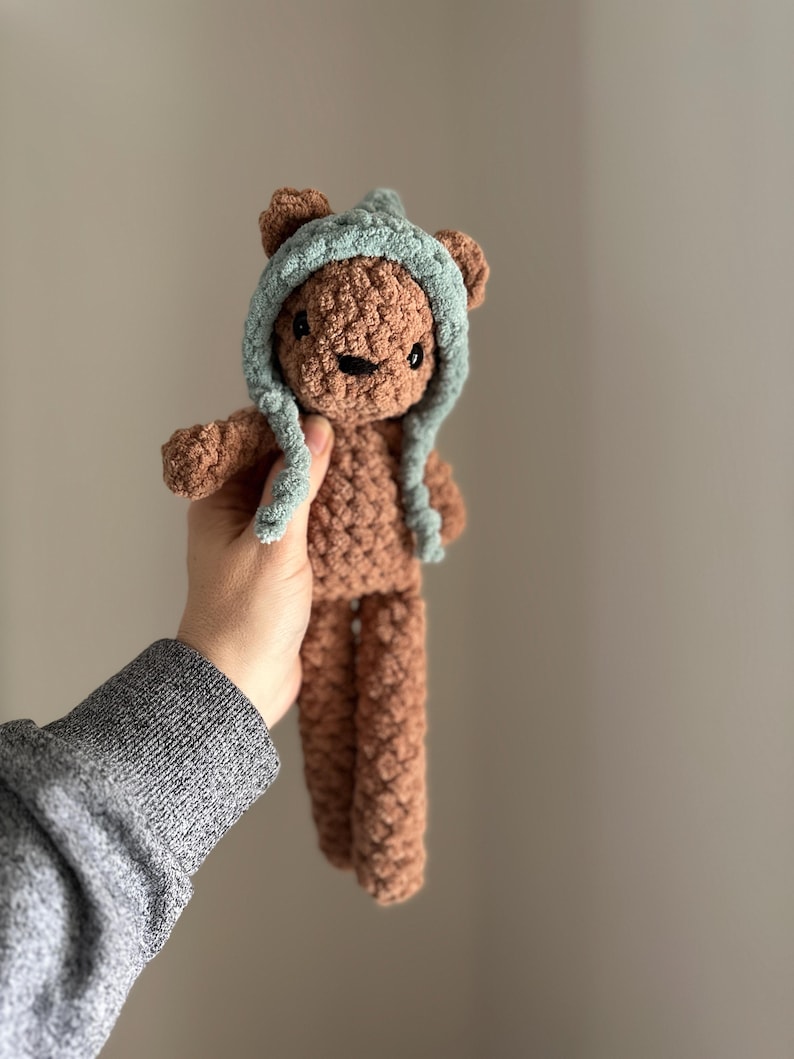 May include: A brown crocheted teddy bear wearing a light blue crocheted hat. The bear has black eyes and a brown nose.