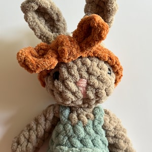 May include: A crocheted plush bunny toy with brown fur, orange ears, and a light blue overall. The bunny has a pink nose and black eyes.