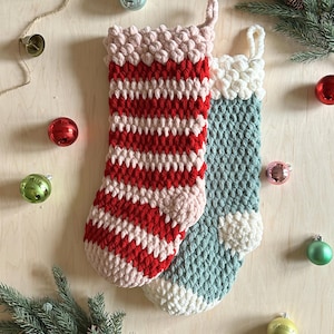 May include: Two crocheted Christmas stockings, one red and pink striped and one teal with white trim. The stockings are on a wooden surface with Christmas decorations.