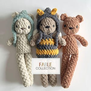 Peut inclure: Trois ours en peluche crochetés, un en beige avec un chapeau bleu, un en marron et un en gris avec un gilet rayé jaune et noir. Les ours sont tous assis sur un fond blanc. Le texte "FABLE COLLECTION" est visible sur l'image.