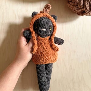 May include: A handmade, plush cat doll in a pumpkin costume. The cat is dark gray with yellow eyes and a white nose. The pumpkin outfit is orange with a stem. The doll is approximately 8 inches tall.