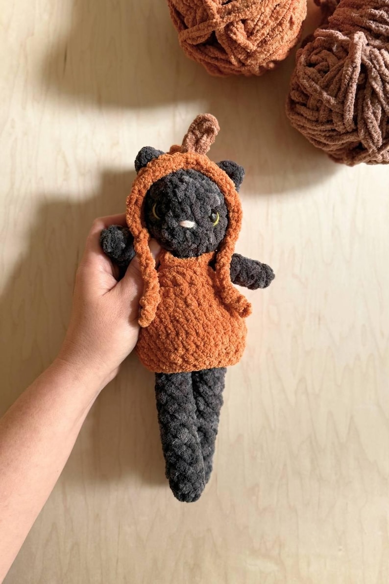 May include: A handmade, plush cat doll in a pumpkin costume. The cat is dark gray with yellow eyes and a white nose. The pumpkin outfit is orange with a stem. The doll is approximately 8 inches tall.