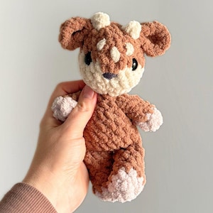 May include: A small, plush, crocheted deer toy made from soft, textured brown and cream yarn. The deer features rounded ears, two small cream-coloured horns on top of its head, dark round eyes, and cream-coloured accents on its paws and face.