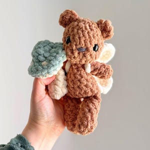May include: A brown crocheted teddy bear plush toy featuring a light blue mushroom cap held in one paw and white wings attached to the back. The bear has black plastic eyes and a small dark nose, crafted from soft, textured yarn material.