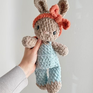 May include: A crochet plush rabbit toy made of soft, chunky beige yarn. The rabbit features long ears, black safety eyes, and is wearing a bright coral-coloured headband with a bow and light blue overalls over its beige body.