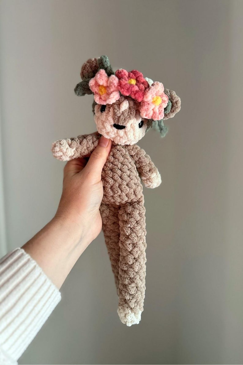 K&ouml;nnte beinhalten: Ein geh&auml;keltes Pl&uuml;schtier in Form eines Rehs mit einer Blumenkrone. Die Krone ist aus rosa und gr&uuml;nem Garn gefertigt und hat verschiedene rosa Blumen.