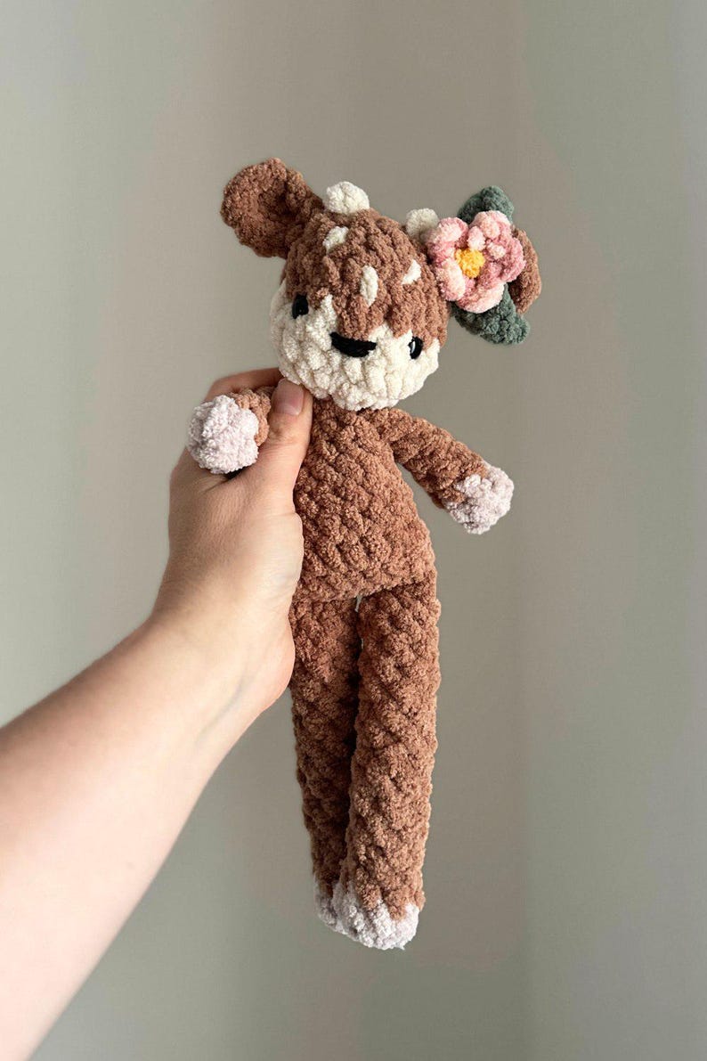K&ouml;nnte beinhalten: Ein braunes geh&auml;keltes Pl&uuml;schtier in Form eines Rehs mit einer rosa Blume auf dem Kopf. Das Reh hat wei&szlig;e Akzente im Gesicht und an den F&uuml;&szlig;en.