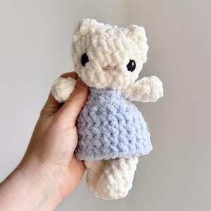 May include: A handmade, plush white cat doll with black eyes and a pink nose. The cat is wearing a light blue dress. The doll is made of soft, textured yarn and has small arms and legs.