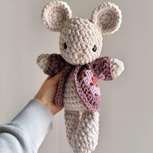 May include: A crocheted stuffed animal resembling a mouse with light beige yarn, large rounded ears, and dark safety eyes. The mouse wears a textured, open-front cardigan in shades of dusty rose and mauve, featuring a small pink heart detail.