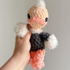 May include: A handmade crochet ostrich doll with a peach-colored head and feet, a gray body, and white fluffy accents. The ostrich has black eyes and a small white tuft on top of its head. The doll is held in a person's hand.