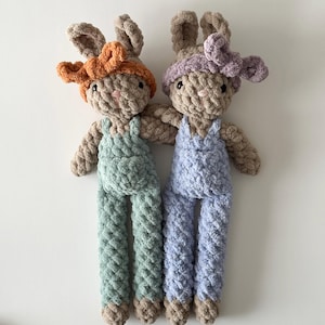 May include: Two crocheted bunny plush toys with long legs and brown fur. One bunny is wearing a light blue overall and a purple headband. The other bunny is wearing a light green overall and an orange headband.