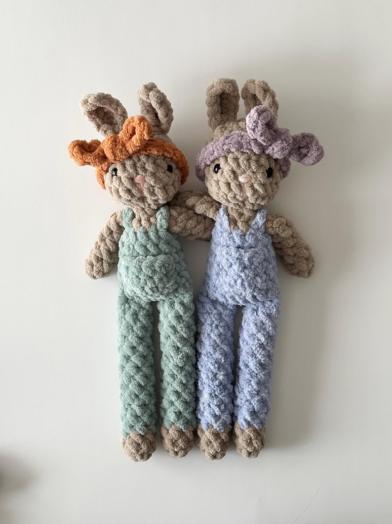 May include: Two crocheted bunny plush toys with long legs and brown fur. One bunny is wearing a light blue overall and a purple headband. The other bunny is wearing a light green overall and an orange headband.