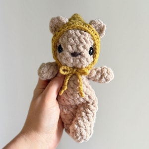 Könnte beinhalten: Ein handgefertigter, beigefarbener Häkelbär mit einer senfgelben Kapuze und Schleife. Der Bär hat schwarze Augen und eine kleine braune Nase. Das Kuscheltier ist weich und kuschelig, perfekt für ein Kinderzimmer oder als Dekoration.