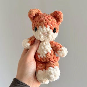 May include: A handmade crochet cat doll in orange and white. The plush toy has a white face, chest, and paws, with orange accents on the ears and body. The cat has black eyes and a pink nose. The doll is soft and cuddly.