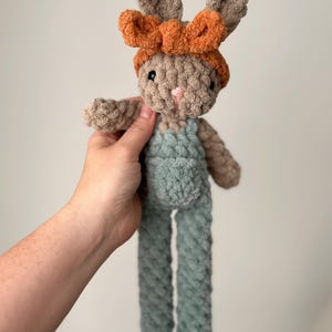 May include: A crocheted plush bunny toy with long legs and a brown and orange headband. The bunny is wearing a light blue overall.