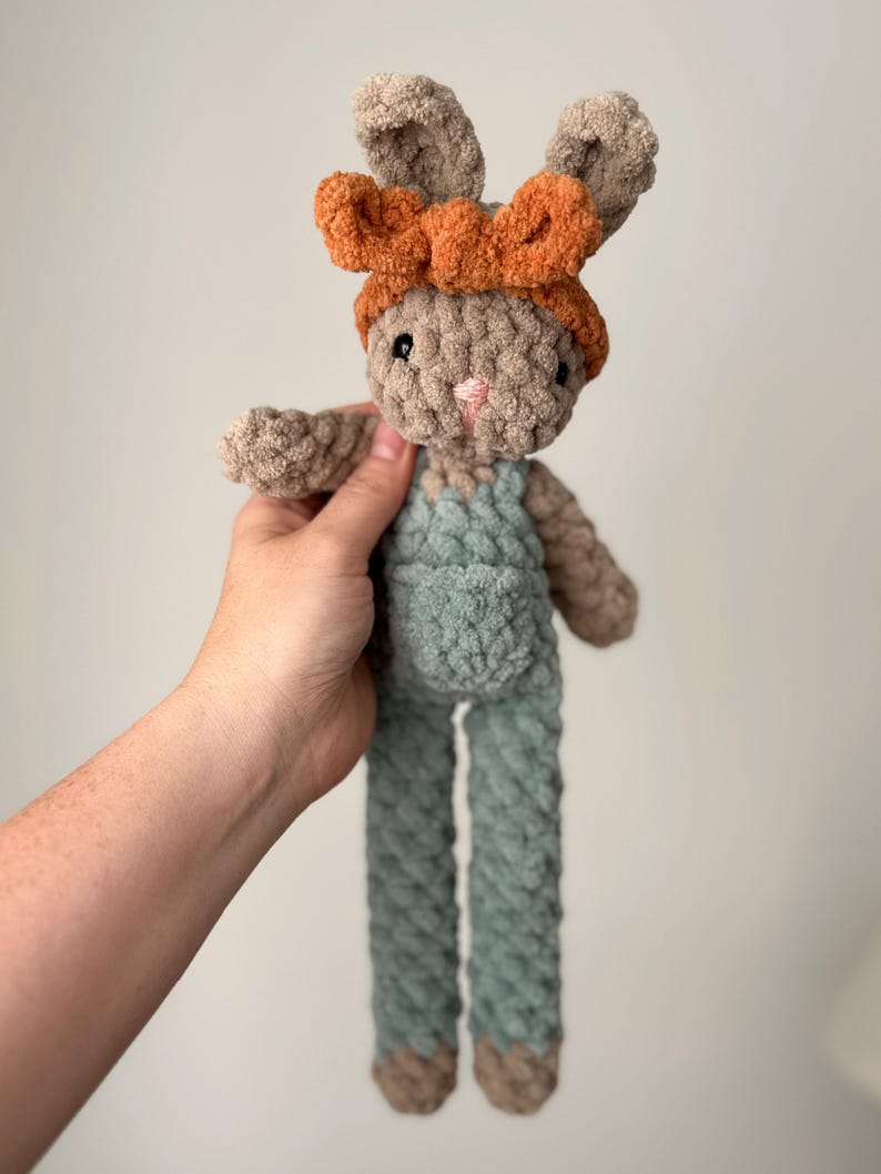 May include: A crocheted plush bunny toy with long legs and a brown and orange headband. The bunny is wearing a light blue overall.
