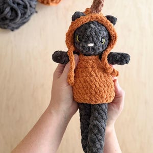 May include: A handmade crochet cat doll in a pumpkin costume. The cat is gray with yellow eyes and a white nose. The pumpkin costume is orange with a brown stem. The doll is held in two hands.