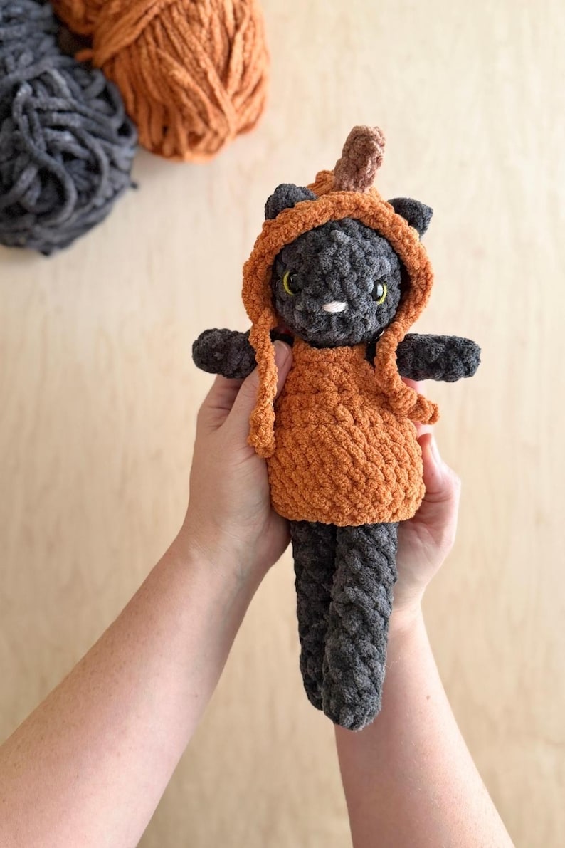 May include: A handmade crochet cat doll in a pumpkin costume. The cat is gray with yellow eyes and a white nose. The pumpkin costume is orange with a brown stem. The doll is held in two hands.