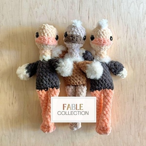 May include: Three handmade crochet ostrich dolls with peach, gray, and brown tones. Each doll has a fluffy white collar, black eyes, and a small white pom-pom on top of its head. The dolls are standing in front of a wooden background. The text "FABLE COLLECTION" is on a white rectangle.