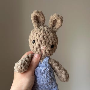 May include: A crocheted plush bunny toy wearing blue dungarees. The bunny has brown fur and black eyes.