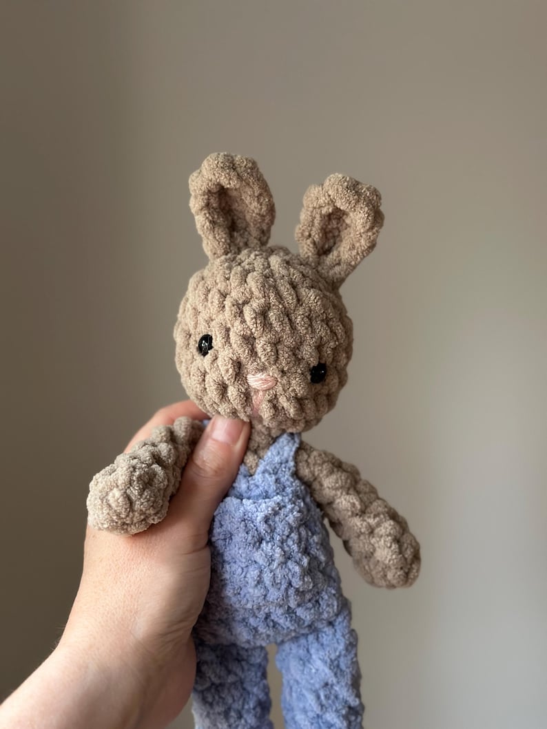 May include: A crocheted plush bunny toy wearing blue dungarees. The bunny has brown fur and black eyes.