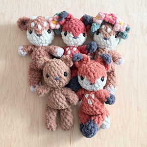 Puede incluir: Una colección de cinco peluches tejidos a mano con hilo suave y texturizado. El grupo incluye tres figuras de ciervos con manchas blancas y detalles florales, un oso marrón sólido y un zorro rojo con patas y orejas de color gris oscuro.