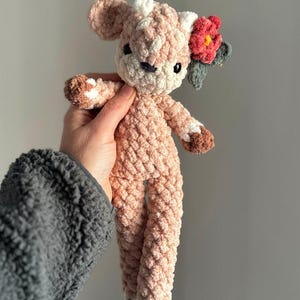 K&ouml;nnte beinhalten: Ein geh&auml;keltes rosa Hirsch-Pl&uuml;schtier mit wei&szlig;en Akzenten und einer roten Blume auf dem Kopf.