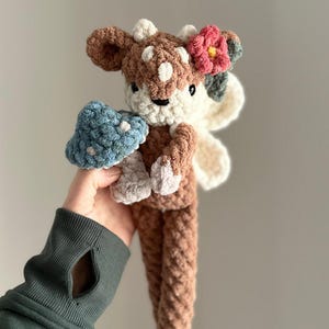 K&ouml;nnte beinhalten: Ein geh&auml;keltes Pl&uuml;schtier in Form eines Rehs mit braunem und wei&szlig;em K&ouml;rper, einer rosa Blume auf dem Kopf und einem blauen Pilz in der Hand.