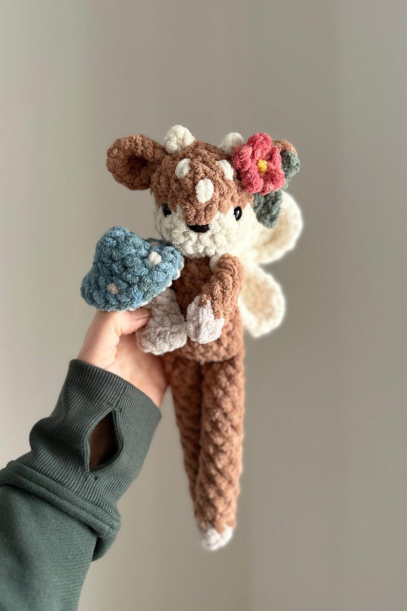 K&ouml;nnte beinhalten: Ein geh&auml;keltes Pl&uuml;schtier in Form eines Rehs mit braunem und wei&szlig;em K&ouml;rper, einer rosa Blume auf dem Kopf und einem blauen Pilz in der Hand.