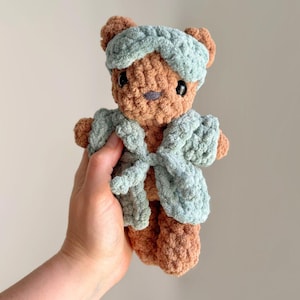 May include: A brown crocheted teddy bear plush toy wearing a light sage green crocheted headband and a matching sage green crocheted robe tied at the front. The bear has dark plastic safety eyes and a small dark grey embroidered nose.