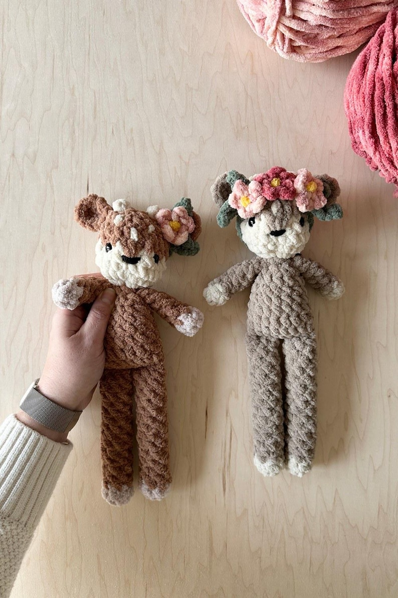 K&ouml;nnte beinhalten: Zwei geh&auml;kelte Pl&uuml;sch-Hirsche mit Blumenkr&auml;nzen. Die Hirsche sind braun und beige mit wei&szlig;en Akzenten. Die Blumenkr&auml;nze sind rosa und gr&uuml;n.